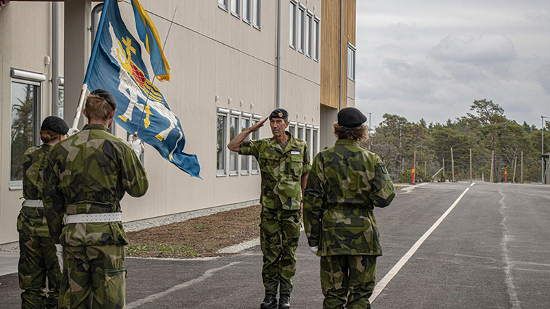 Överbefälhavaren och två militärer hälsar med en honnör utanför en byggnad i trä. 