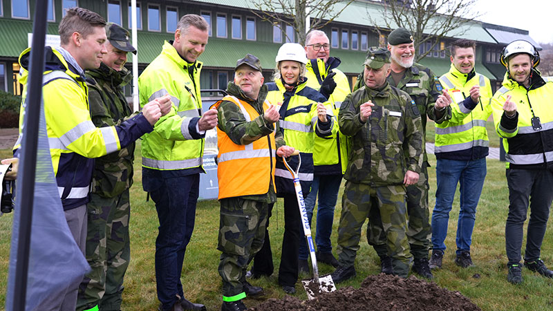 Entreprenörer och militärer i reflexvästar står på rad med spadar och knäpper fingrarna. 