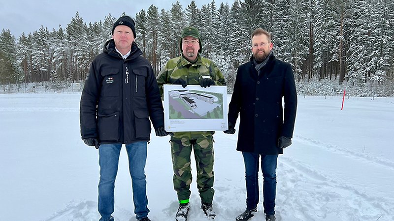 Tre män står i ett snöigt landskap med snötäckta granar bakom sig. De står vända mot kameran och tillsammans håller de upp en illustration som visar nya byggnader.